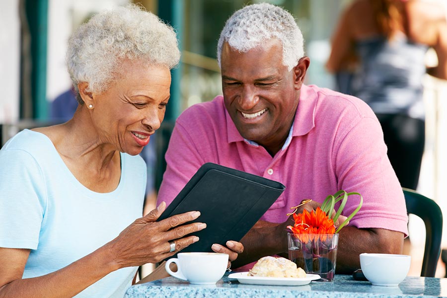 Blog - Senior Couple Reads a Tablet Seated at an Outdoor Table Enjoying Breakfast and Coffee