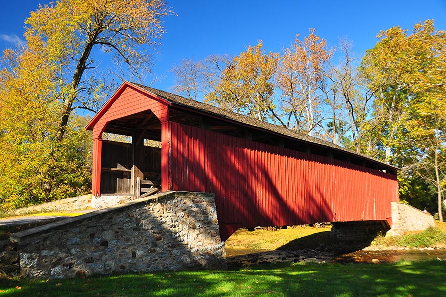 Lancaster, PA - a Red Covered Bridge in Pennsylvania Surrounded by Green Grass and Fall Foliage, a Stream Passing Underneath the Bridge