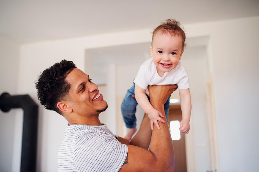 Personal Insurance - Young Father Flies His Toddler Son Through the Air in Their Home, Both Smiling